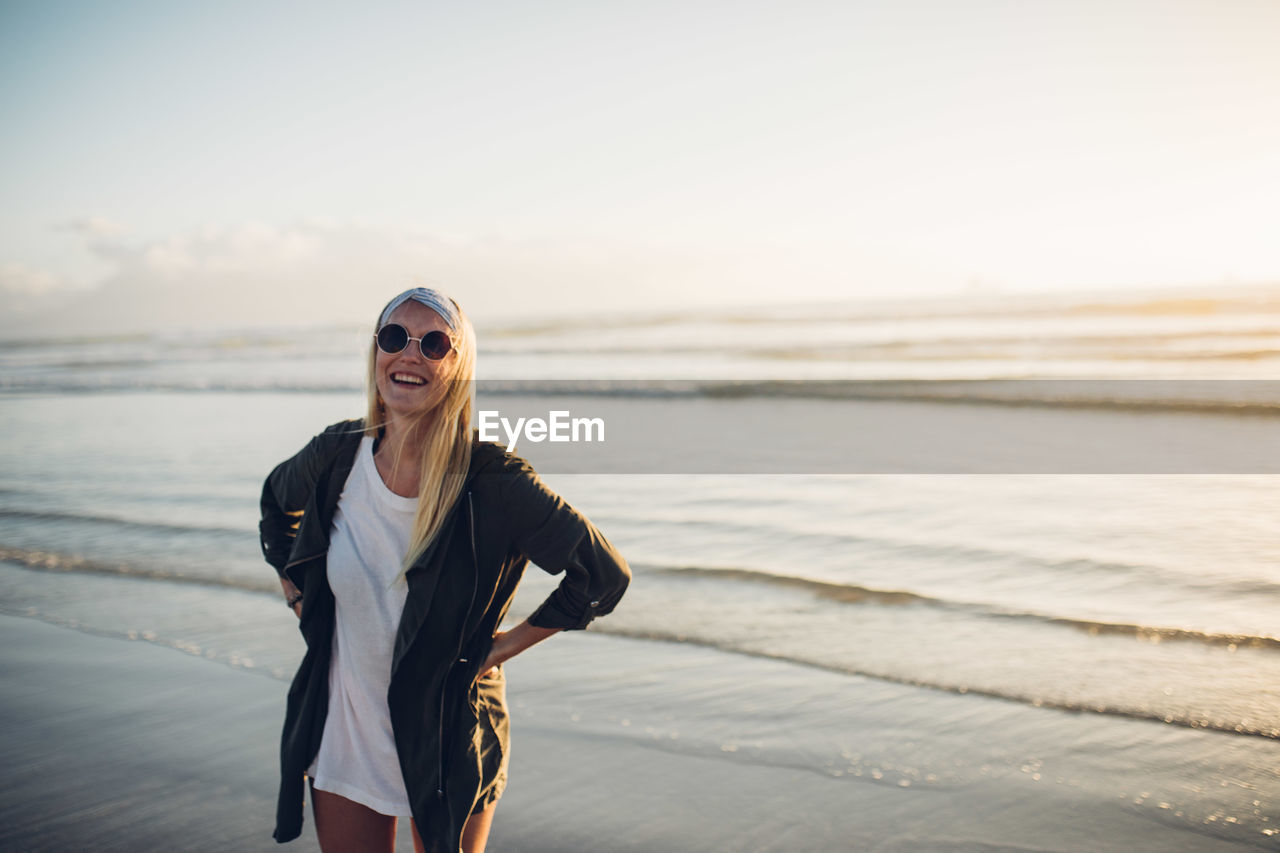 PORTRAIT OF HAPPY YOUNG WOMAN STANDING AT BEACH