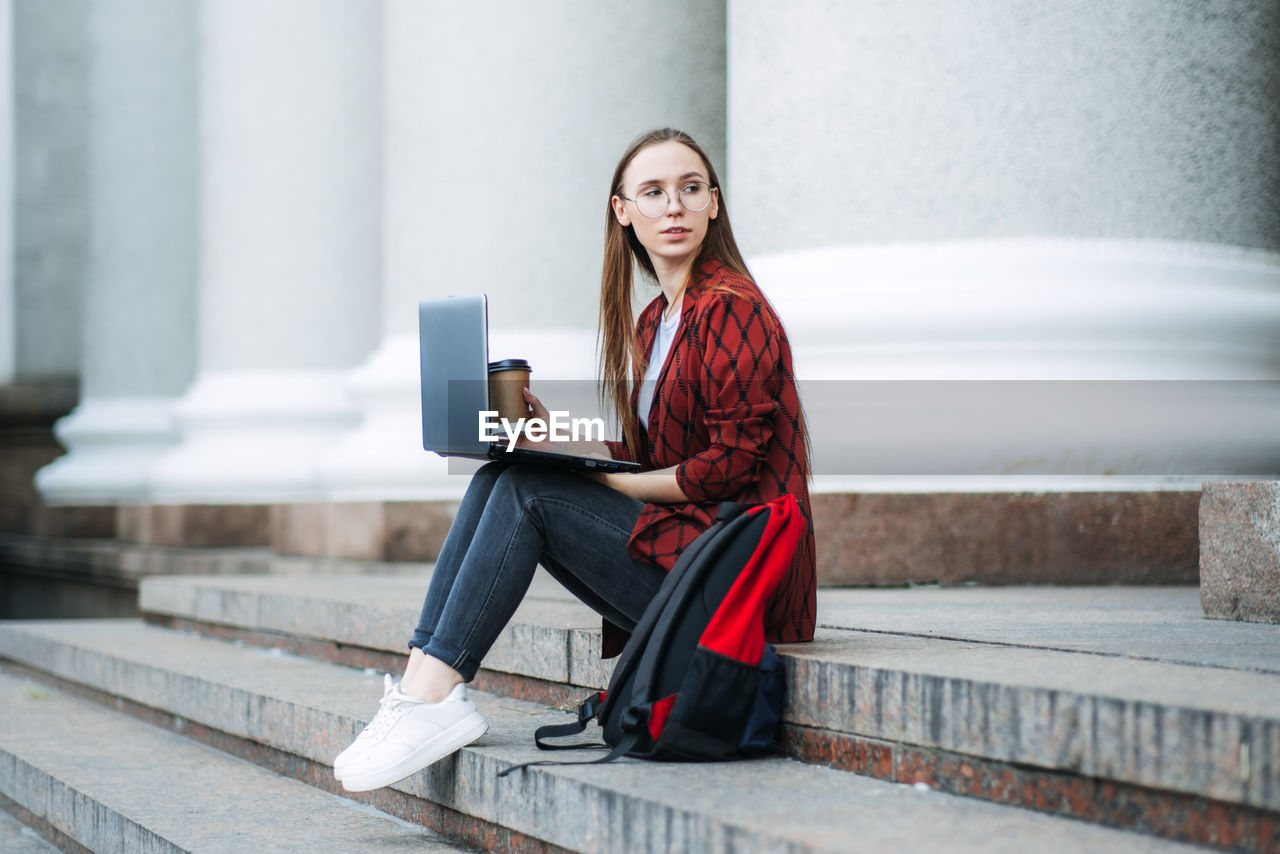 Outdoor portrait of female student with coffee cup and laptop. girl student has coffee break after