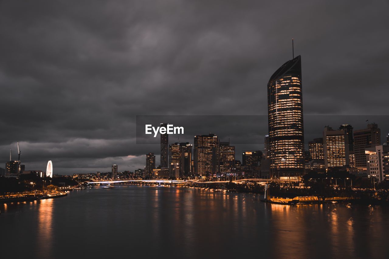 Illuminated city at waterfront against cloudy sky