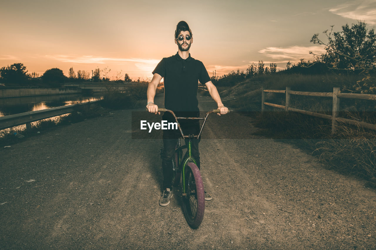 Portrait of young man riding bicycle while standing against sky during sunset