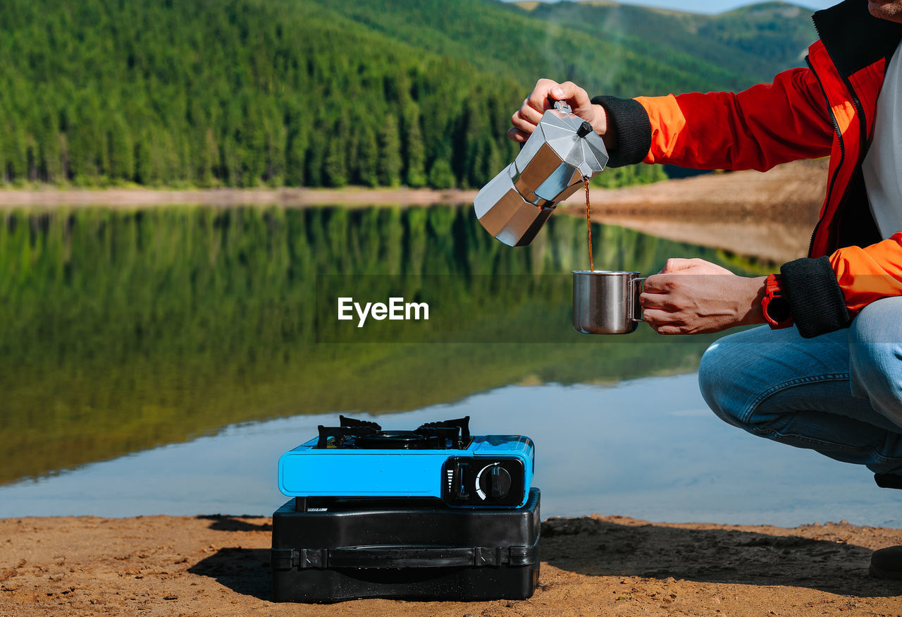 Man holding coffee drink by the camping stove against the lake