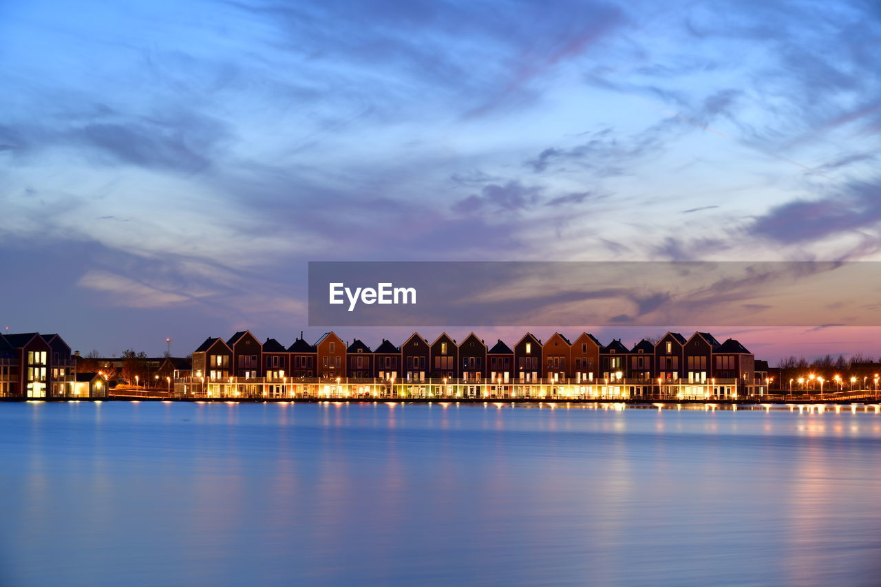 Illuminated buildings by lake against sky during sunset