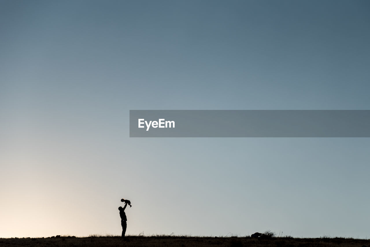 Silhouette father with child standing on field against clear sky during sunset