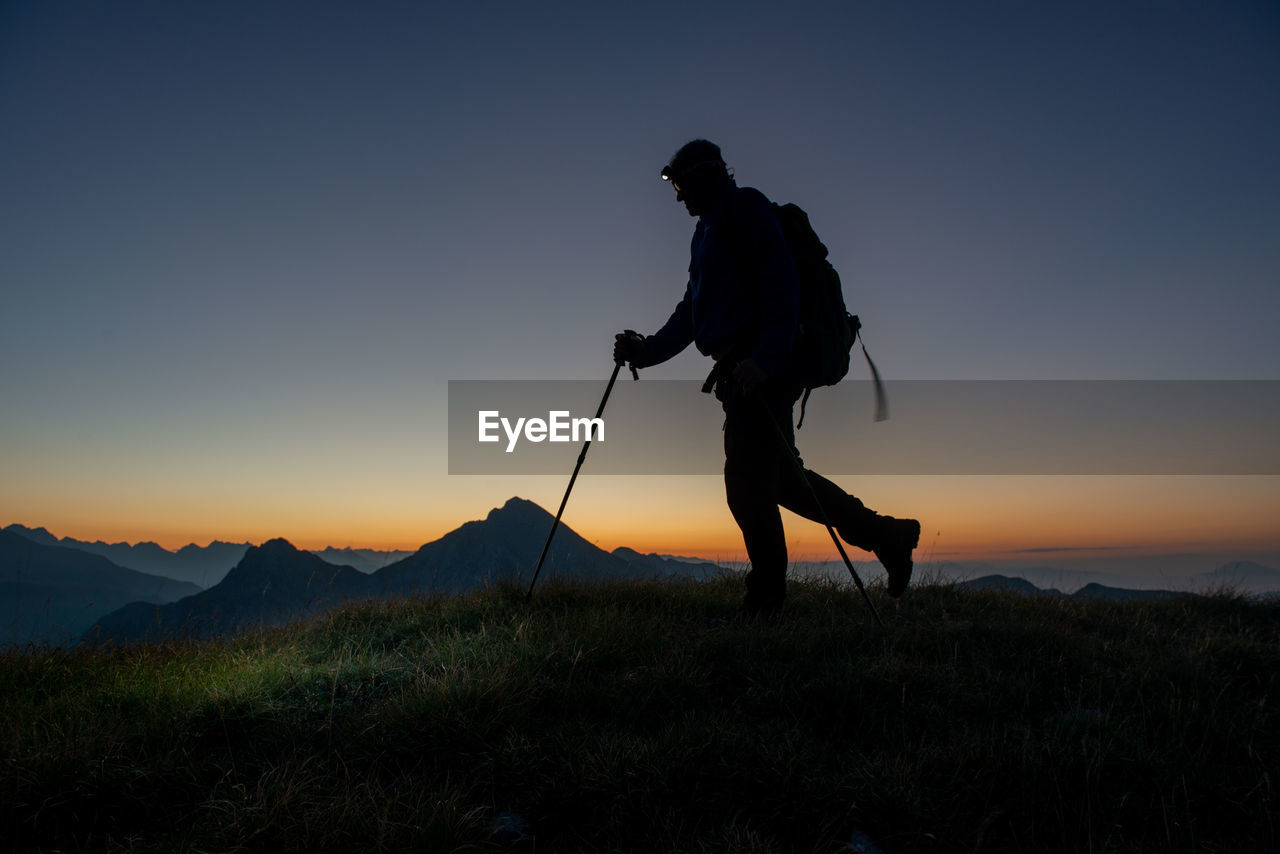sky, silhouette, mountain, activity, one person, adult, full length, nature, leisure activity, sunset, horizon, men, hiking, sports, beauty in nature, person, landscape, scenics - nature, walking, mountain range, golf, dawn, standing, environment, grass, lifestyles, hiking equipment, land, adventure, sun, golf club, outdoors, skiing, side view, back lit, holding, travel, copy space, motion