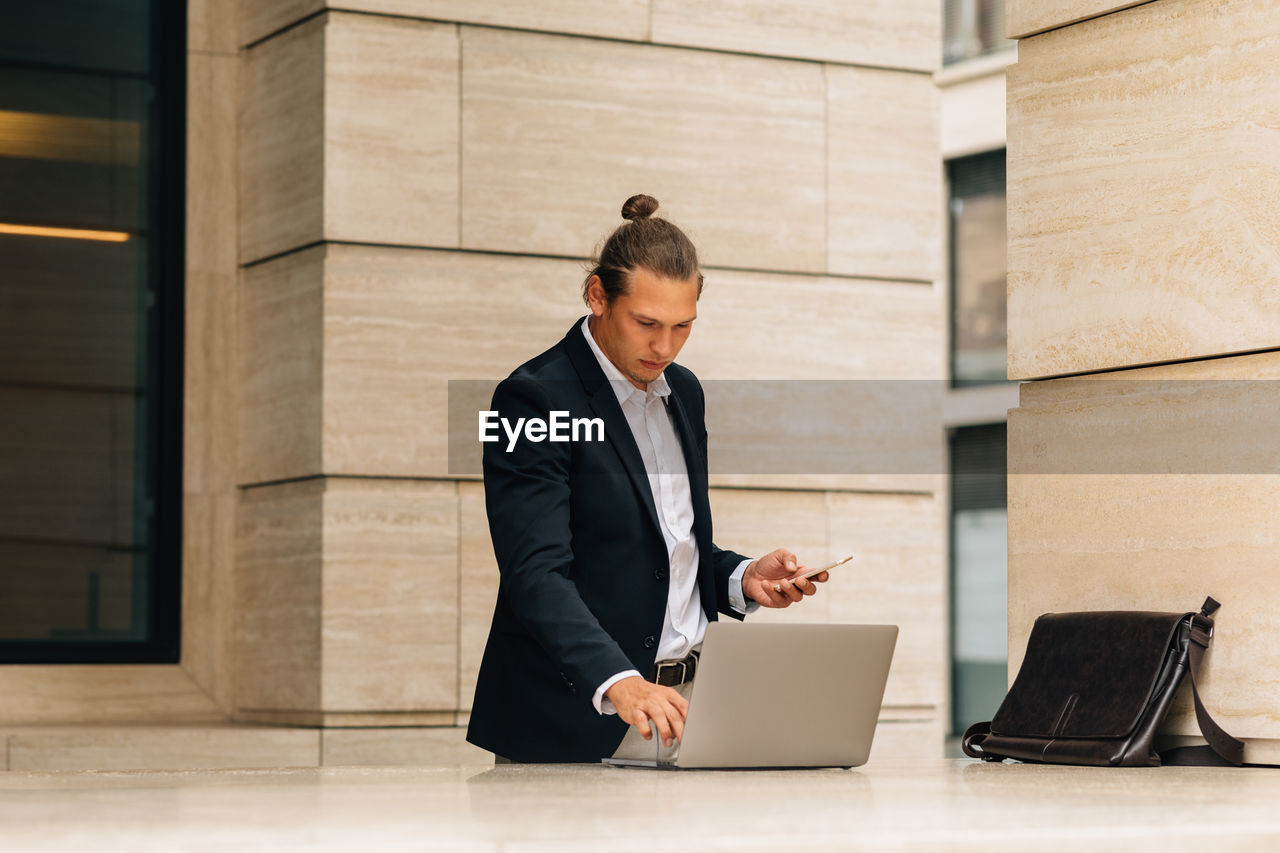 Businessman using laptop while holding smart phone against building