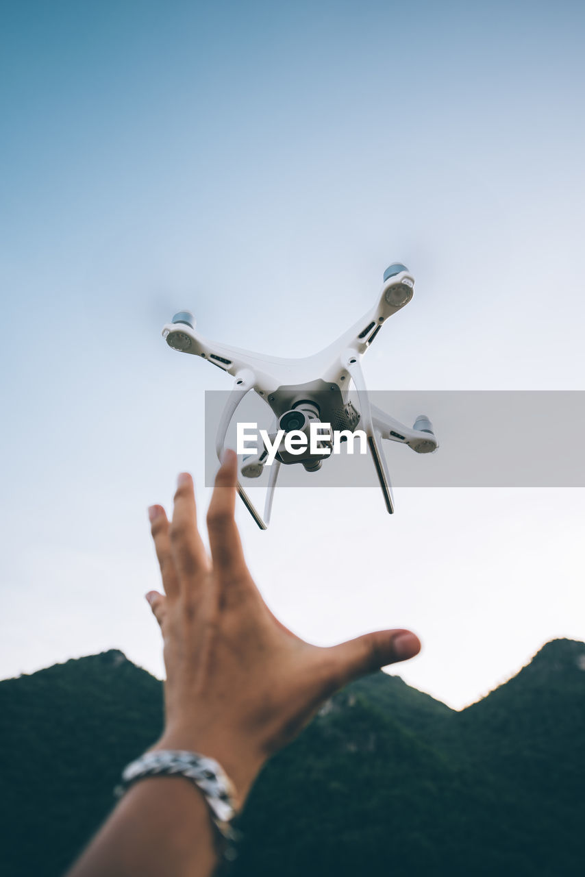 Cropped image of hand holding drone flying against sky