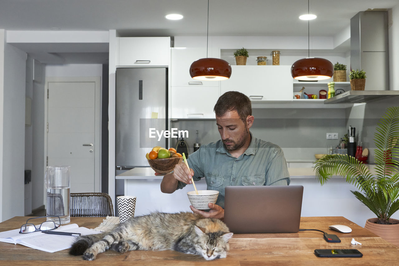 Businessman eating noodles while cat sleeping at dining table