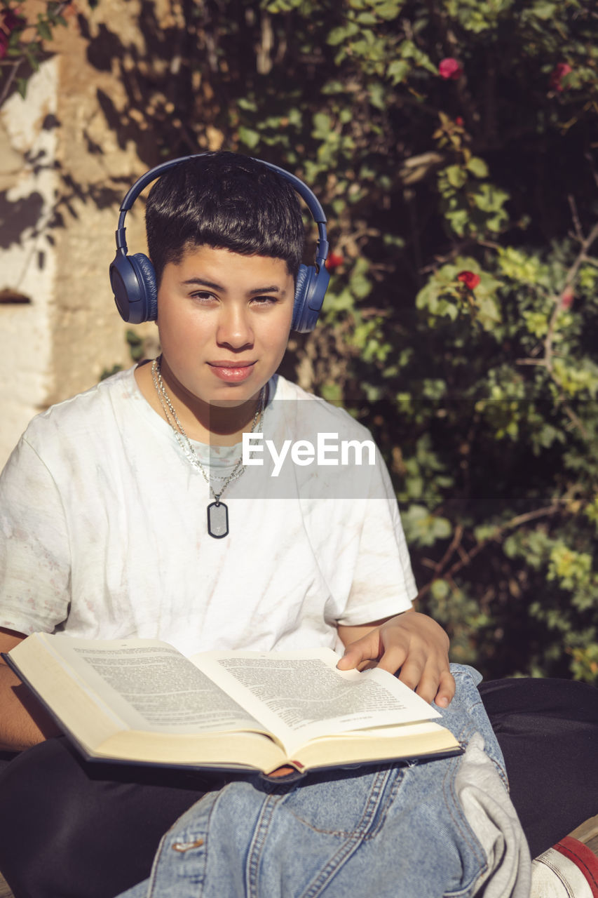 one person, headphones, music, sitting, listening, publication, book, person, reading, child, casual clothing, activity, young adult, lifestyles, leisure activity, clothing, adult, education, arts culture and entertainment, relaxation, spring, learning, student, three quarter length, teenager, front view, portrait, men, childhood, technology, human face, paper, holding, studying, nature, looking, outdoors, serious, adolescence, women