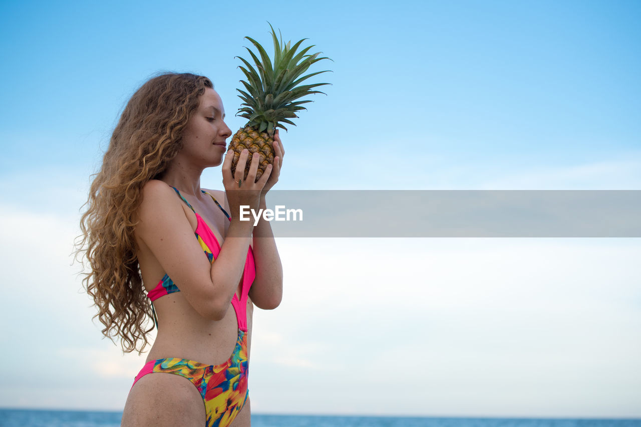Young woman holding pineapple standing against sky