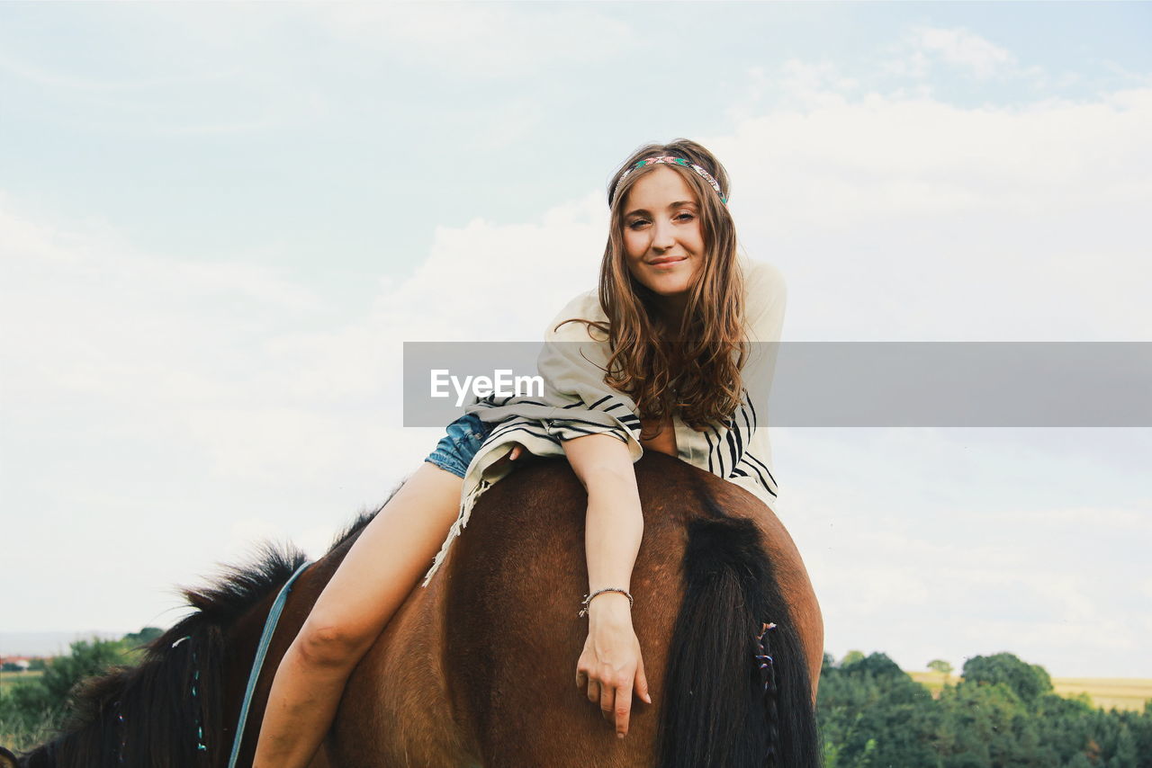 Portrait of beautiful smiling young woman sitting on brown horse on field