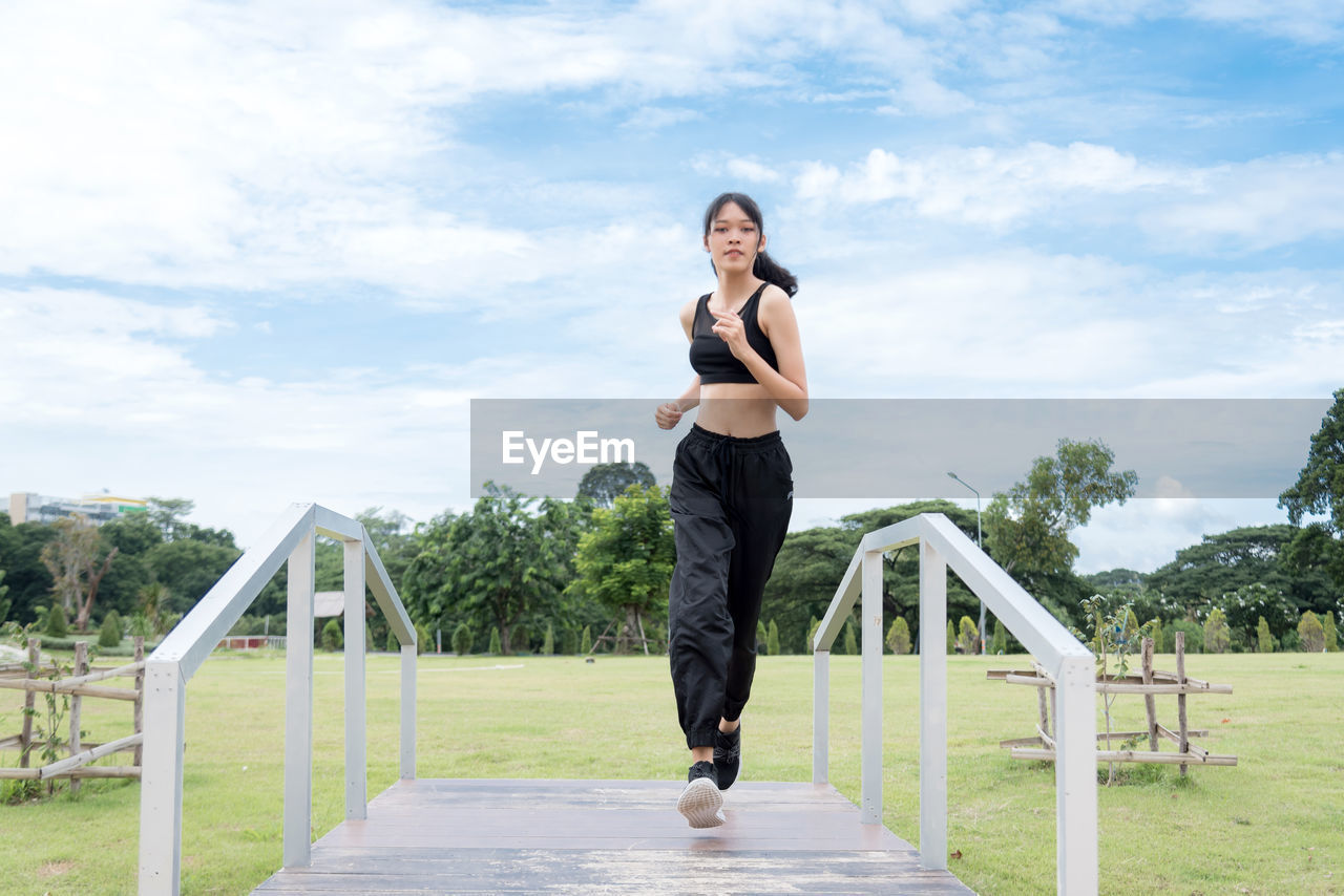 Portrait of woman running on footbridge against sky in park