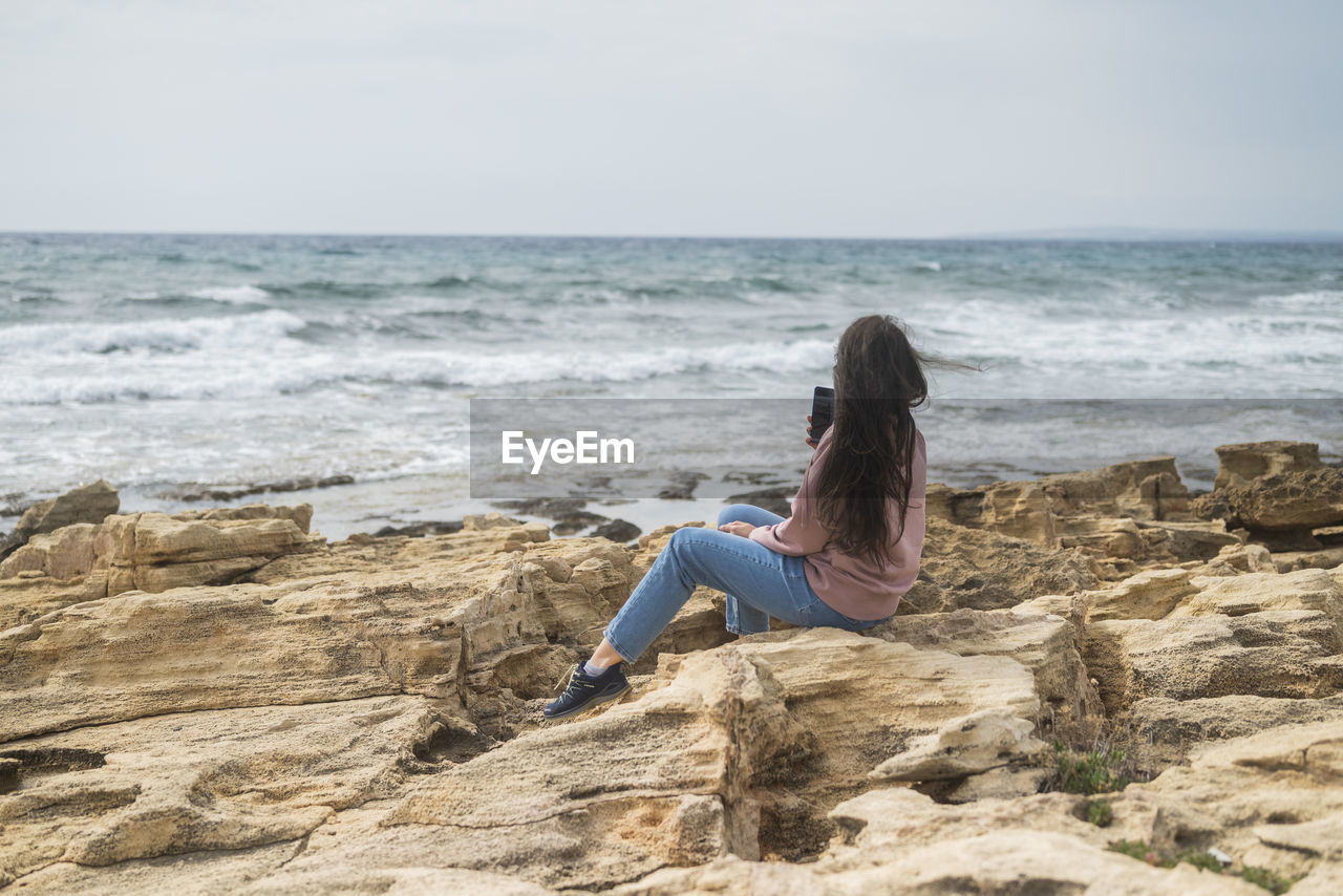 side view of woman sitting on rock at beach against sky