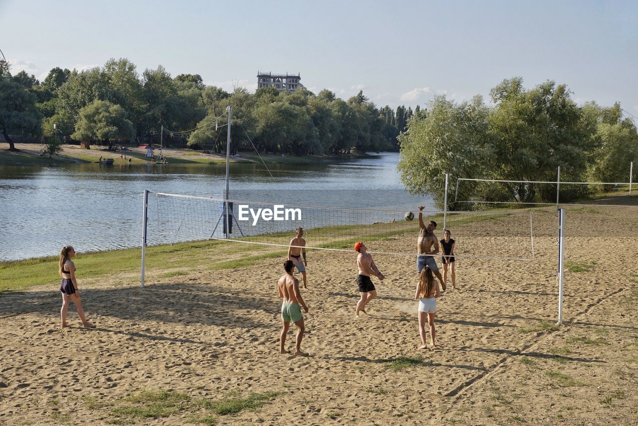 Friends playing volleyball on sand by lake against sky