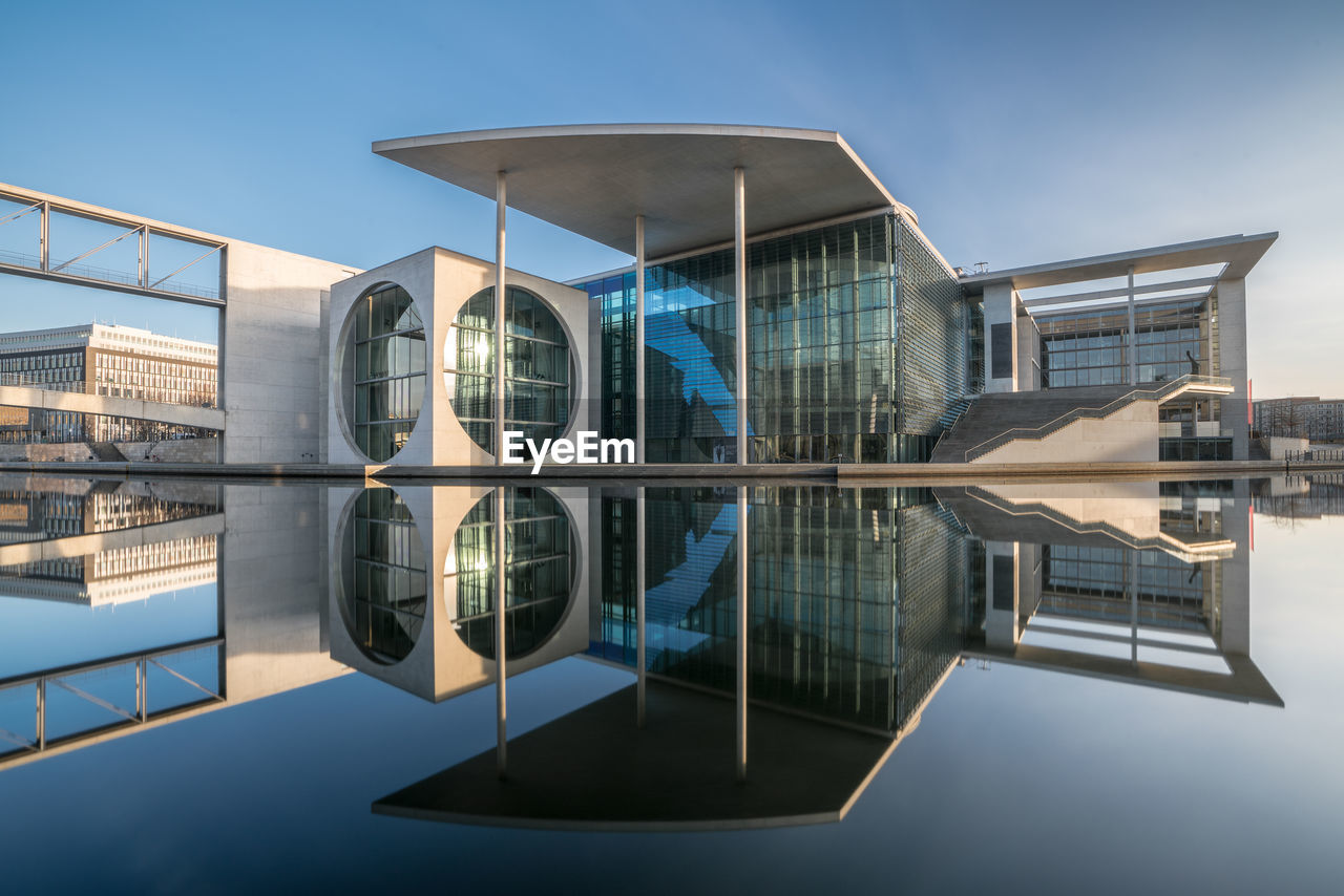 Modern building reflecting on water against clear blue sky during sunny day