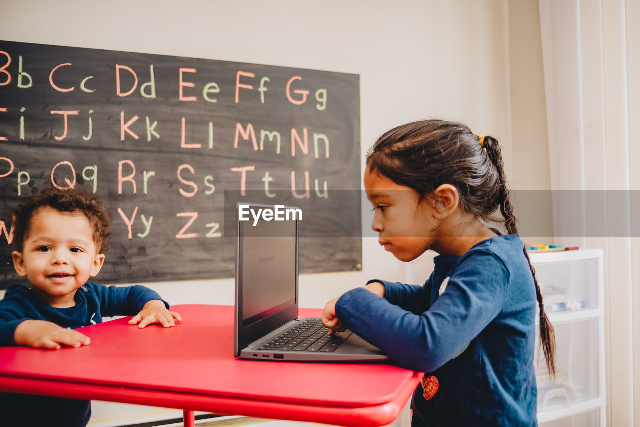 Baby boy standing by table while sister studying over laptop at home