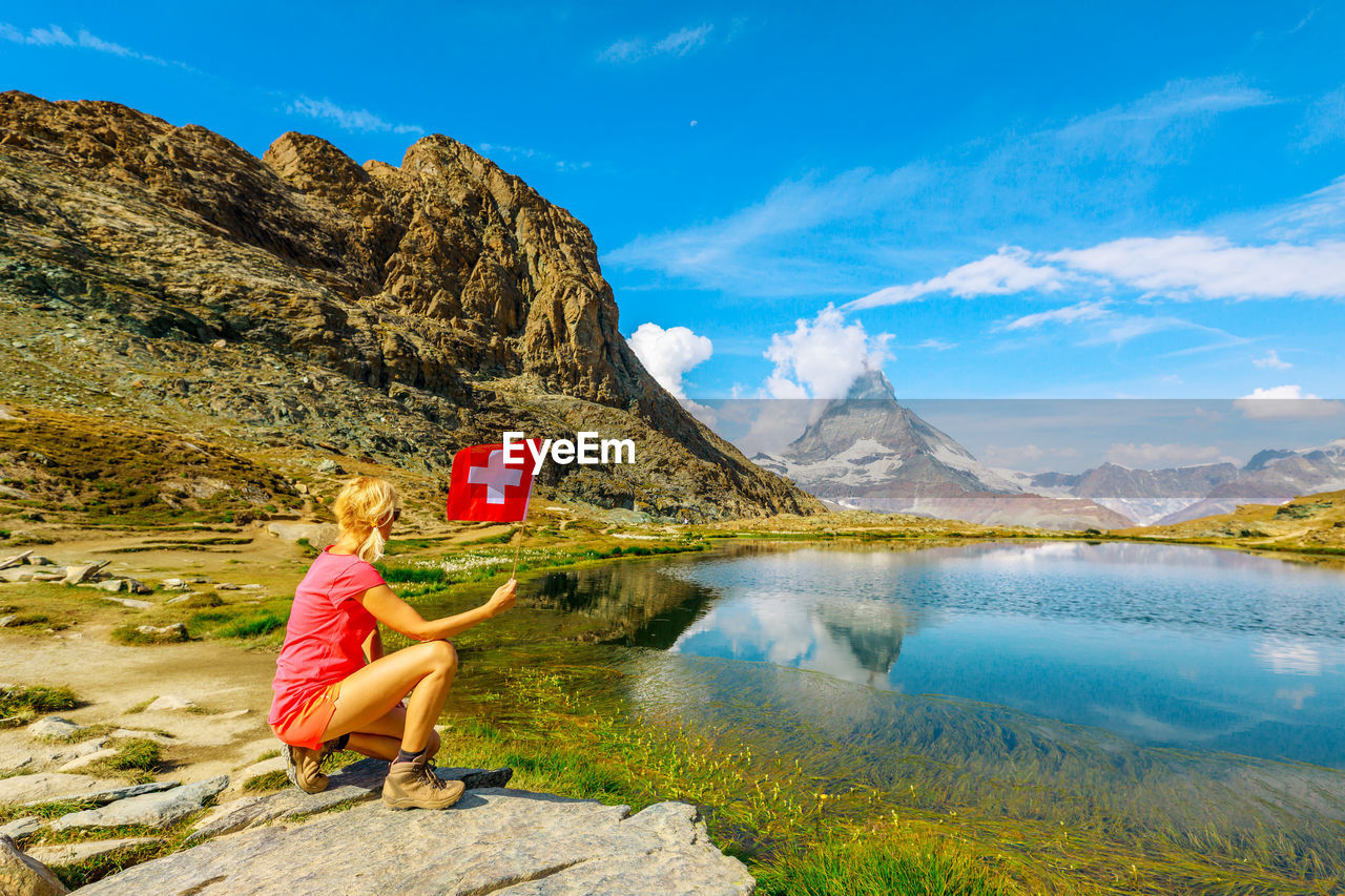 Rear view of woman crouching on rock while holding flag by lake against sky