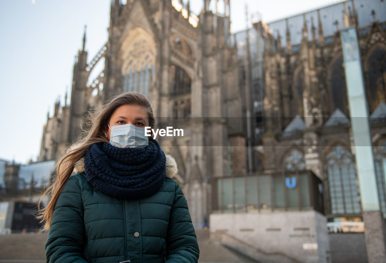 Portrait of young woman wearing surgical mask standing by church outdoors