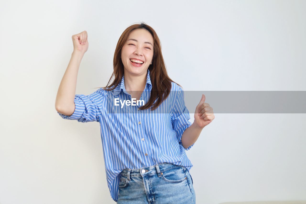 smiling, one person, happiness, adult, emotion, women, indoors, portrait, casual clothing, studio shot, cheerful, positive emotion, young adult, front view, standing, looking at camera, copy space, photo shoot, teeth, smile, blue, waist up, long hair, finger, sleeve, gesturing, relaxation, clothing, enjoyment, arm, communication, female, success, hand, lifestyles, brown hair, white background, joy, hairstyle, limb, button down shirt, person, teenager, jeans, arms raised, business, laughing, human limb