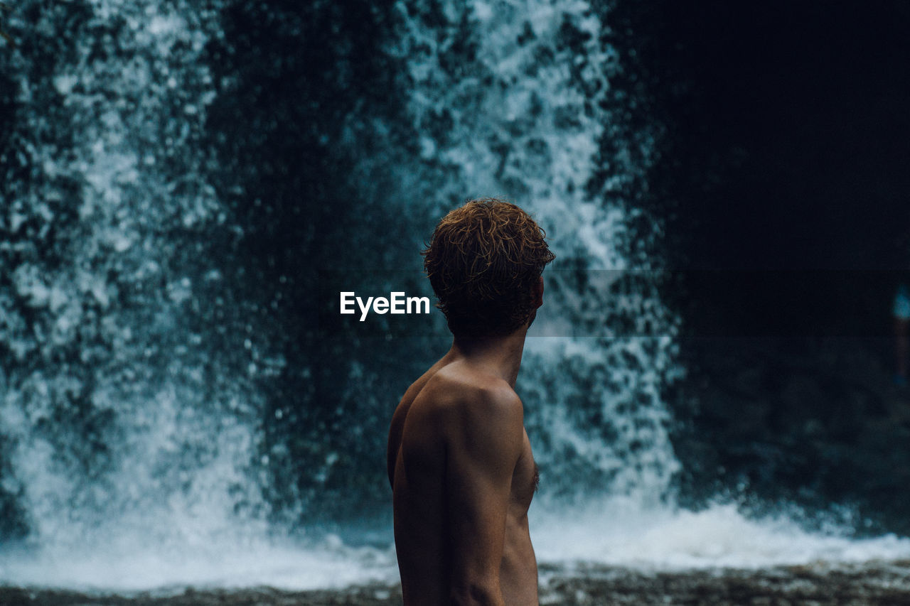 REAR VIEW OF SHIRTLESS MAN LOOKING AT SEA AGAINST WATERFALL