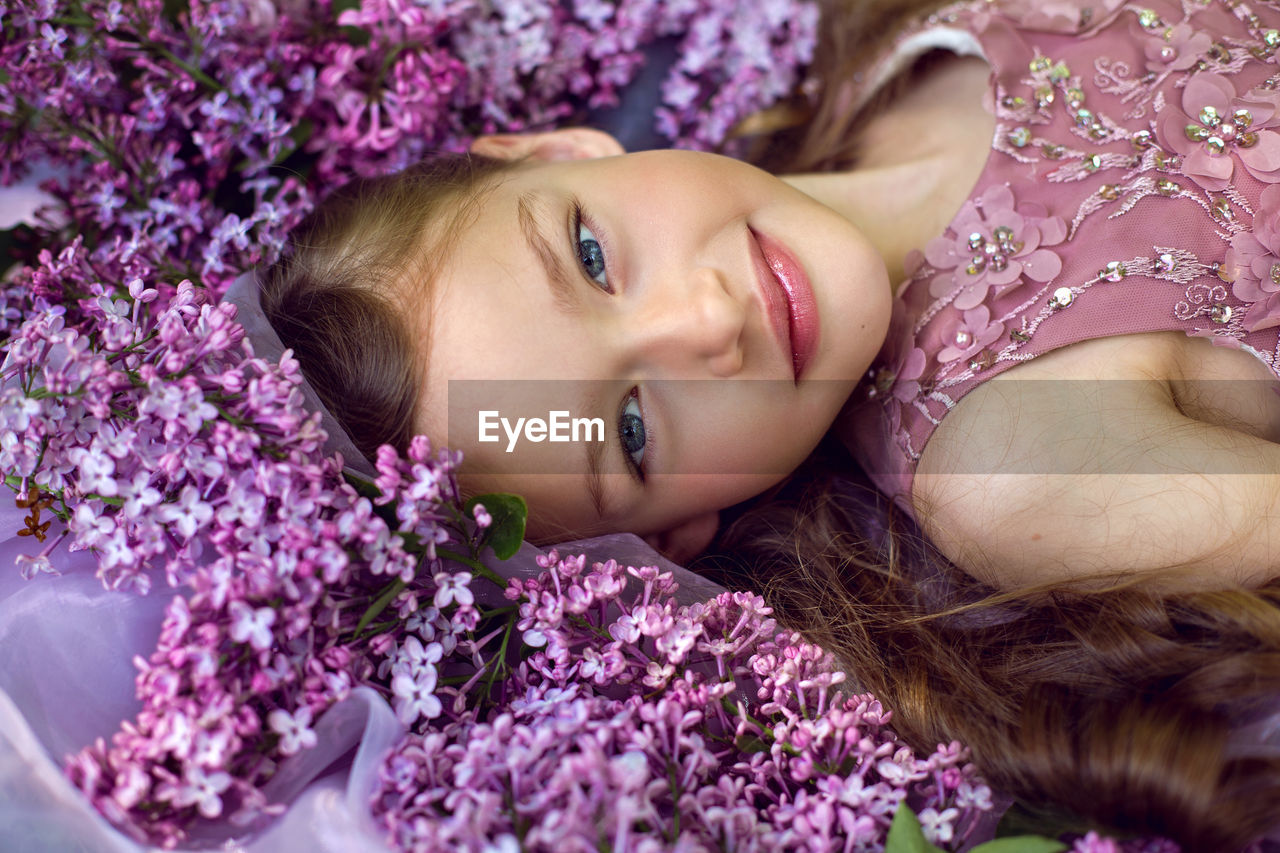 Child girl in a purple floral dress lies on the ground among lilacs on a veil in spring