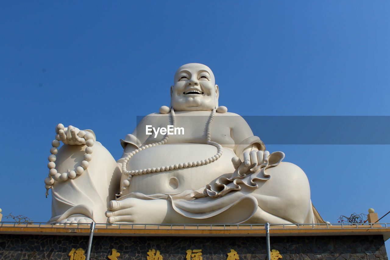 Low angle view of giant laughing buddha against clear blue sky