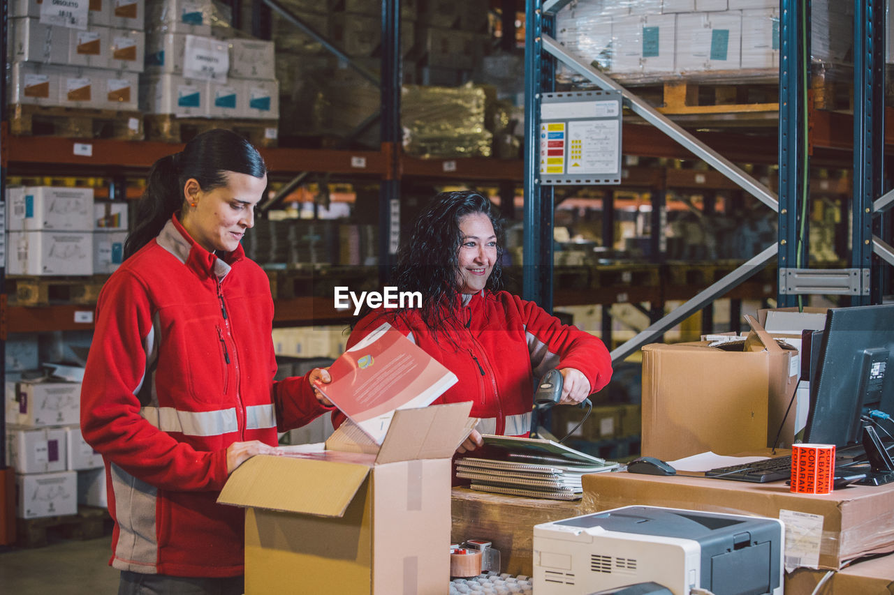 Pleased female employees in red uniform searching packages and collecting parcels in big modern warehouse