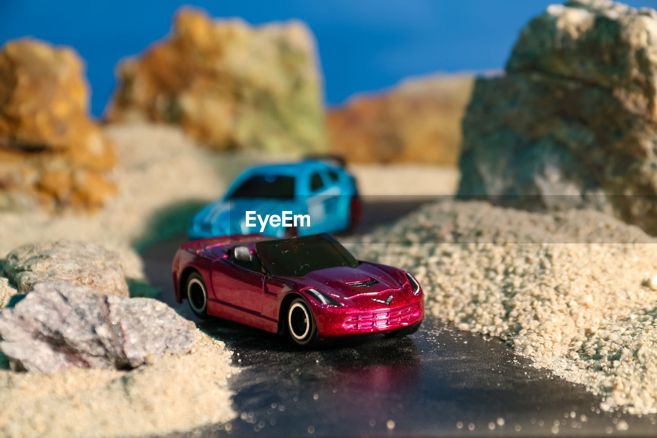 car, vehicle, land vehicle, rock, motor vehicle, transportation, nature, mode of transportation, blue, land, sky, travel, automobile, red, no people, selective focus, toy car, sunlight, focus on foreground, day, wheel, beach, outdoors, water, sunny