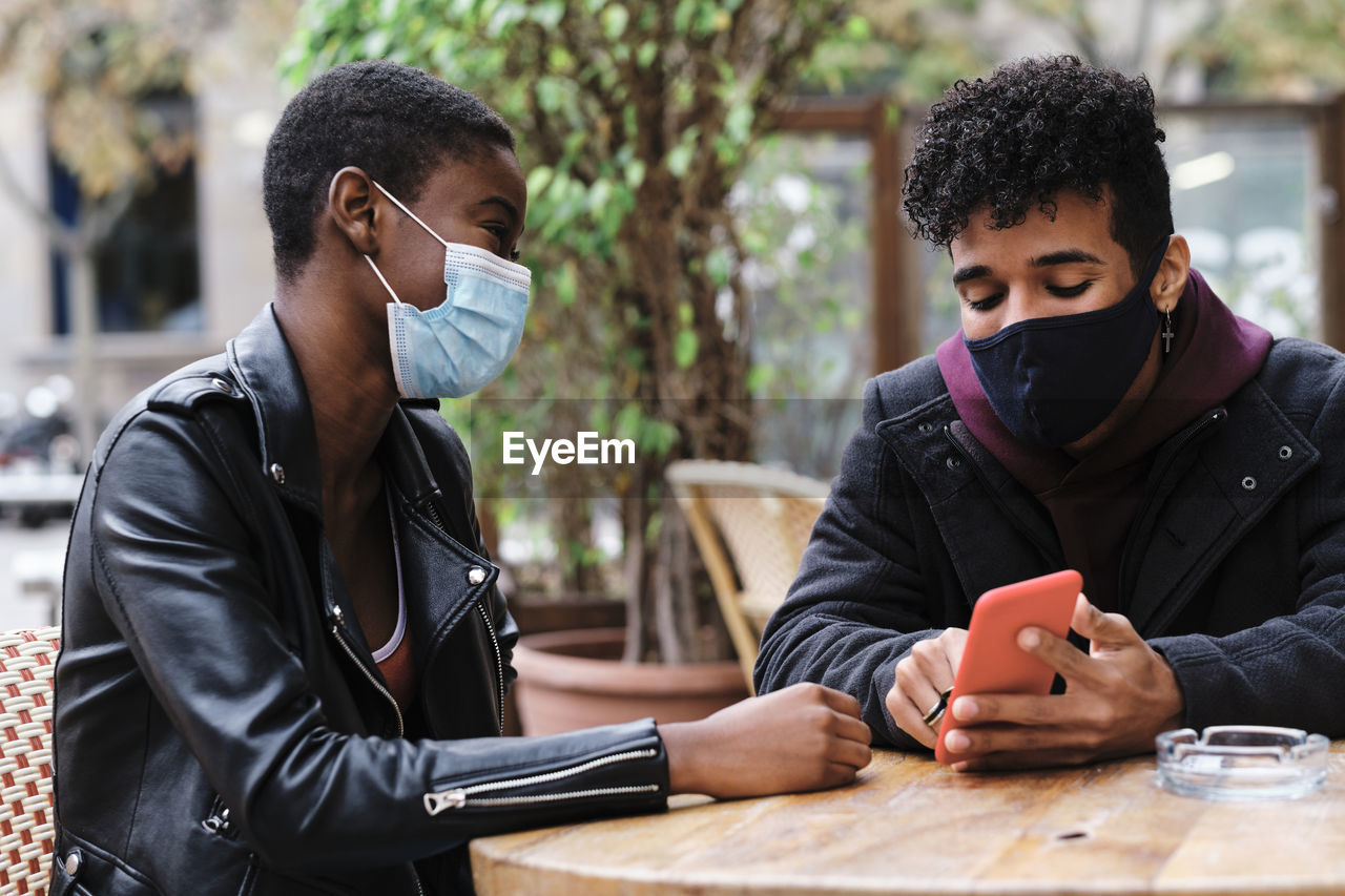 Friends wearing protective face mask using mobile phone while sitting at sidewalk cafe