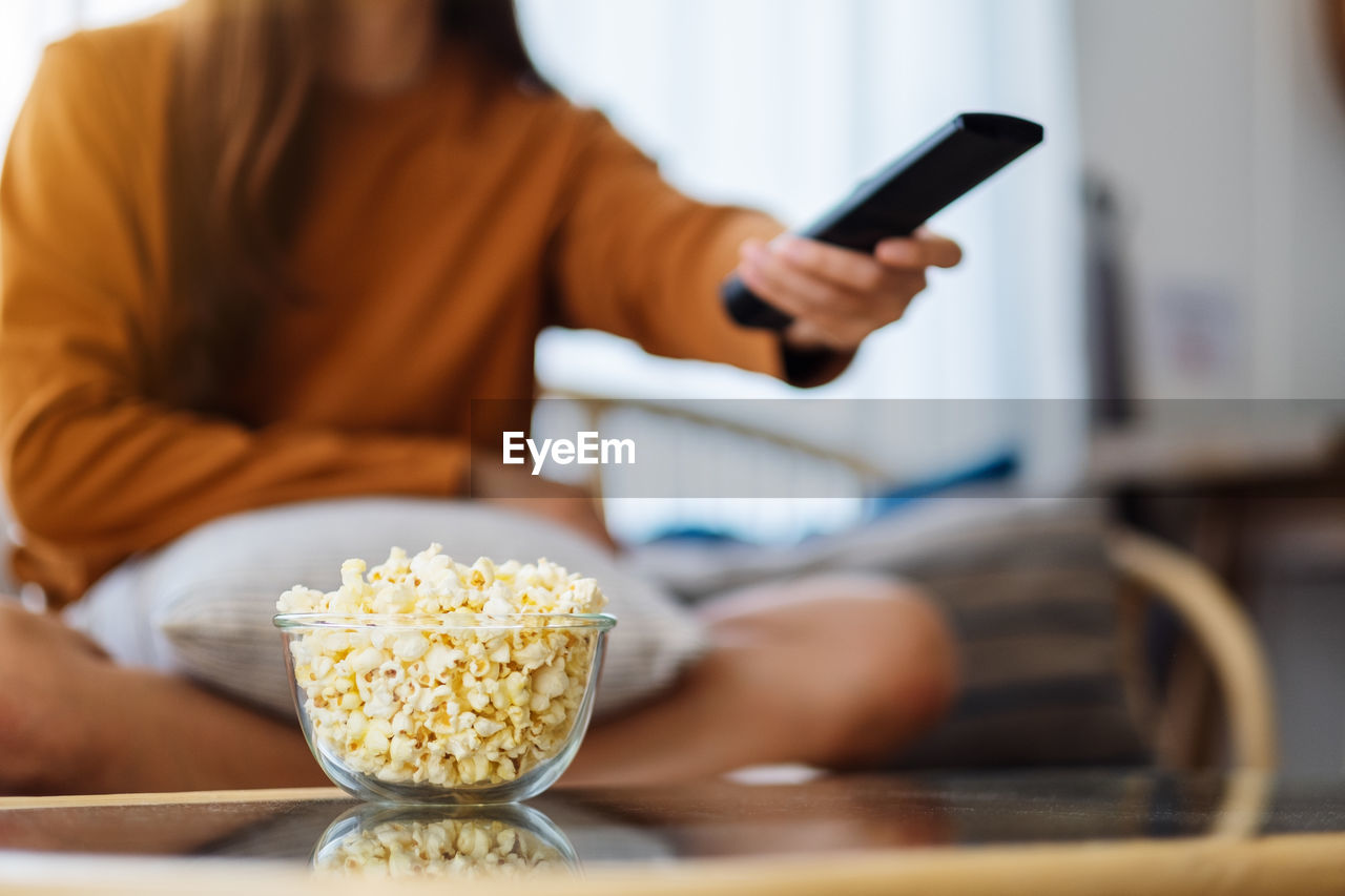 A woman eating pop corn and searching channel with remote control to watch tv while sitting on sofa