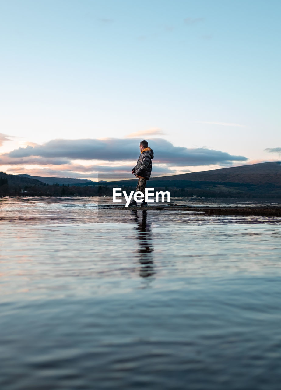 Side view of boy standing by lake against sky