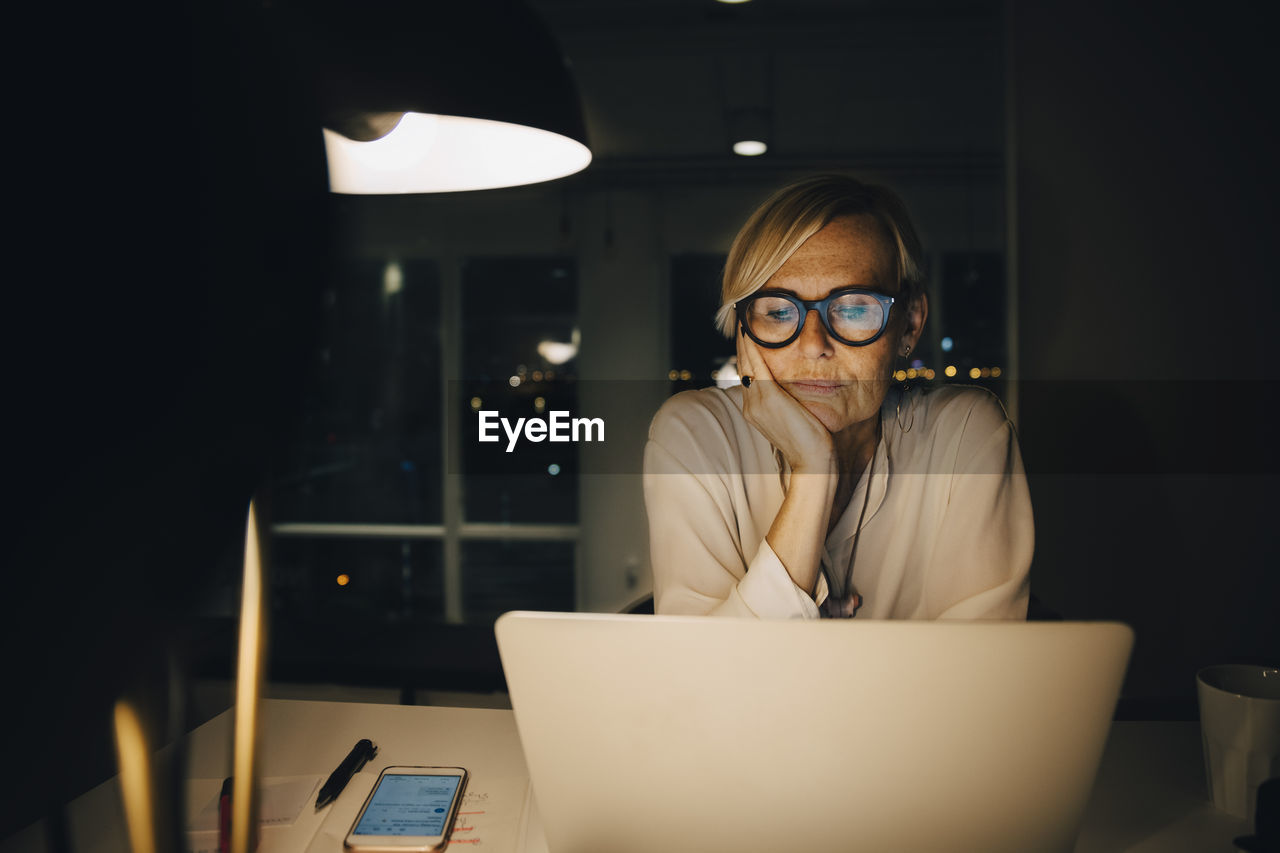 Tired mature female professional staring at laptop while sitting with hand on chin working late in office