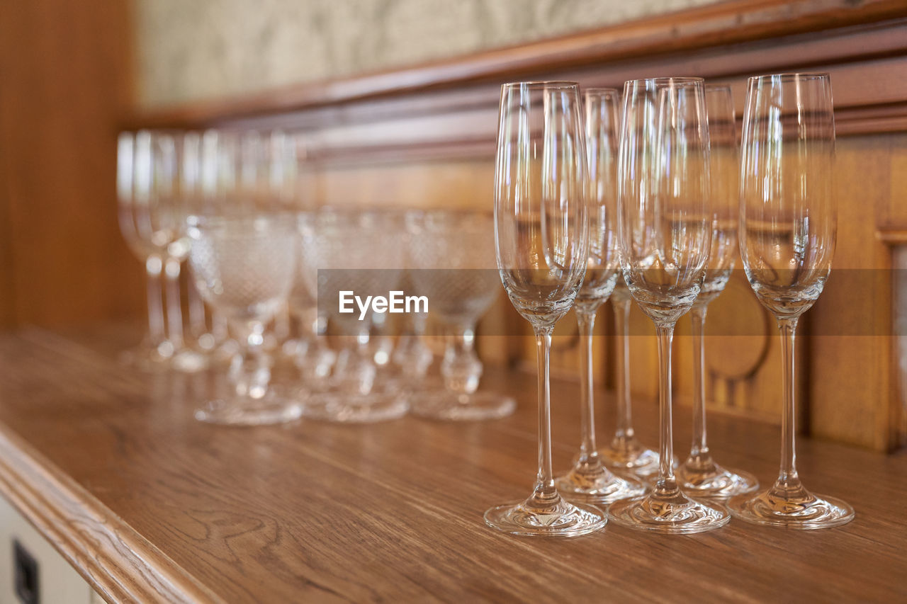 glass, household equipment, food and drink, wine glass, drinking glass, table, indoors, no people, wood, drink, luxury, alcohol, wealth, refreshment, elegance, furniture, business, restaurant, setting, place setting, arrangement, wine, stemware, dining table, focus on foreground, champagne flute, in a row, still life, bar, selective focus, empty, group of objects, domestic room, tableware, order, celebration, decoration