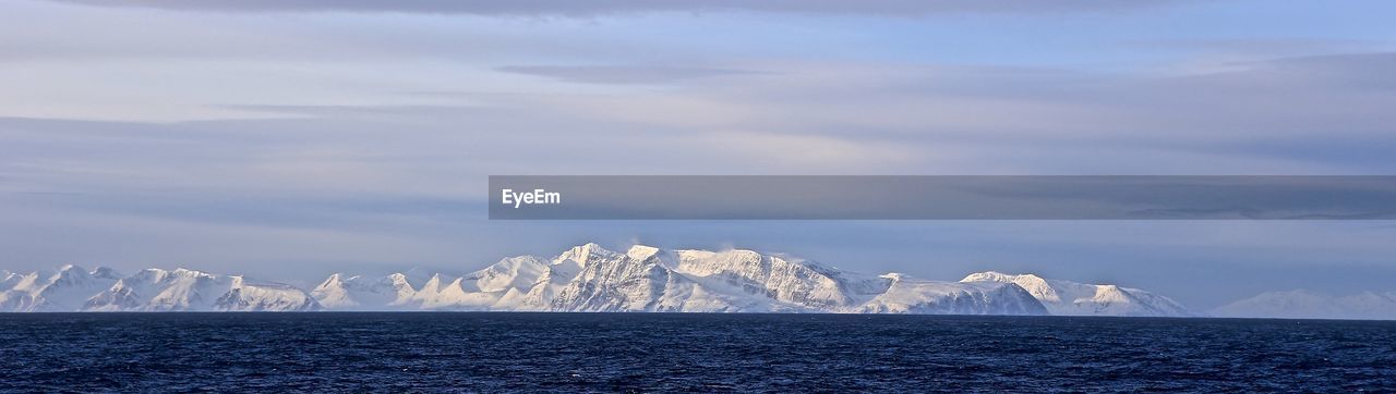 Panoramic view of snowcapped mountains in front of sea against cloudy sky