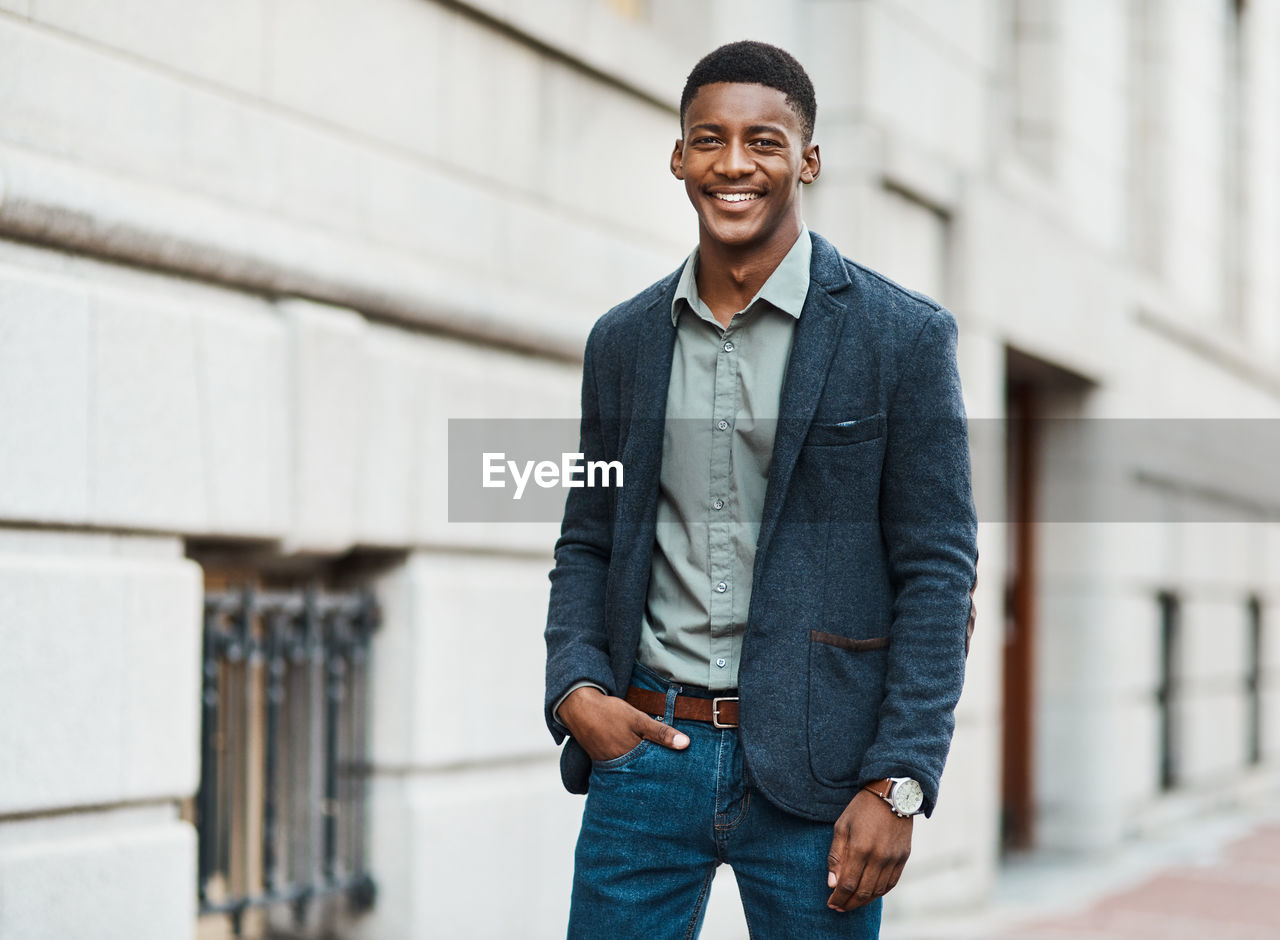 architecture, one person, adult, men, city, smiling, happiness, standing, spring, denim, casual clothing, city life, business, hands in pockets, jeans, portrait, copy space, looking, cheerful, businessman, young adult, clothing, front view, emotion, outerwear, lifestyles, person, building exterior, day, built structure, shirt, three quarter length, technology, teeth, smile, outdoors, street, business finance and industry, wireless technology, blue, beard, blazer, fashion, cool attitude, portable information device, looking at camera, relaxation, mobile phone, facial hair, smartphone, walking, enjoyment