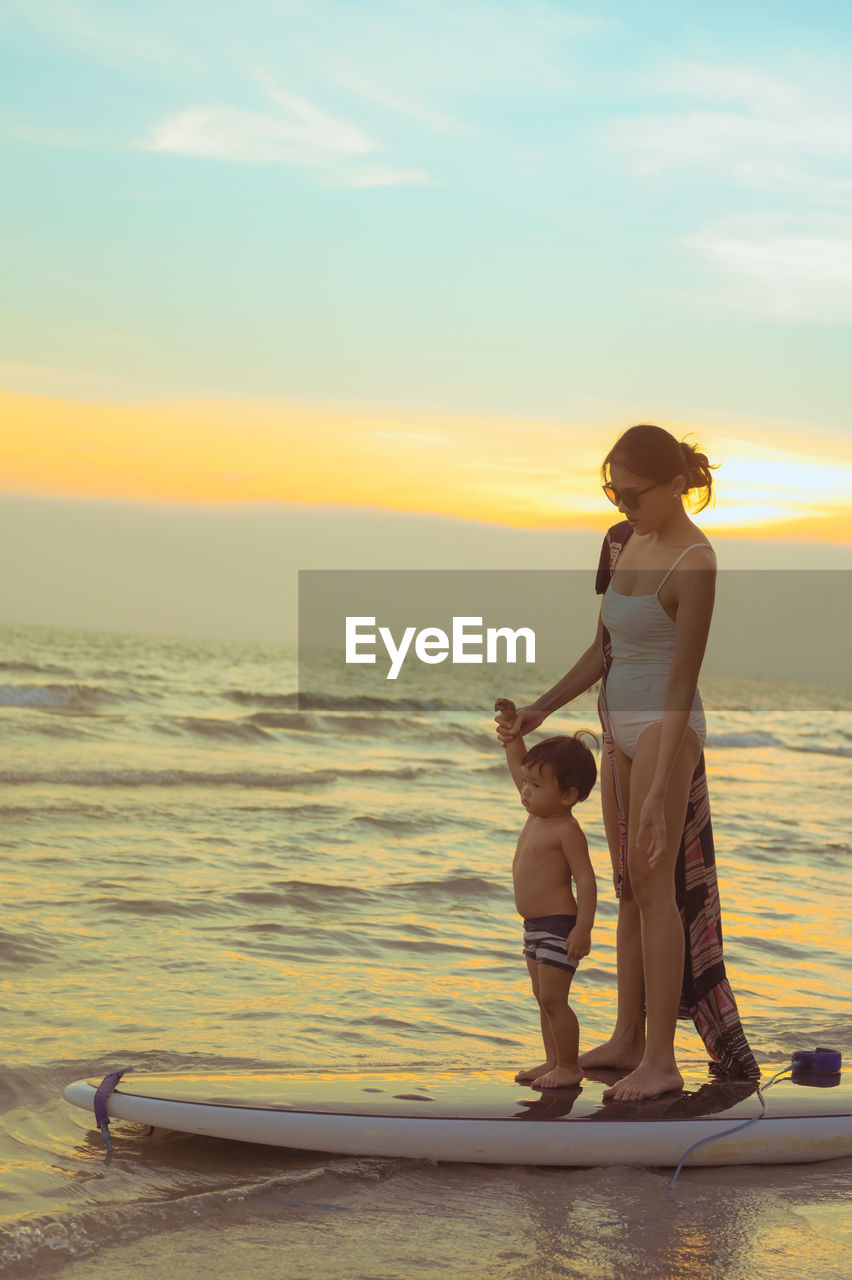 Woman standing on surf board against sky during sunset