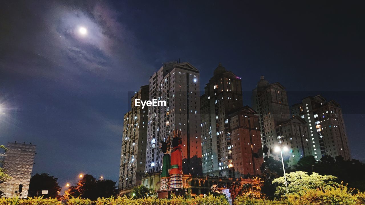 LOW ANGLE VIEW OF ILLUMINATED STREET AMIDST BUILDINGS AGAINST SKY