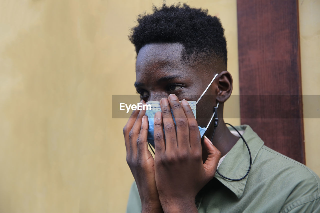 Portrait of young black skin man with face mask and hands cover on its standing against wooden door.