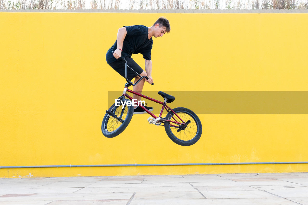 Full body side view male in casual clothes jumping on bmx bicycle against sunlit concrete colorful wall yellow background on summer weekend day on street