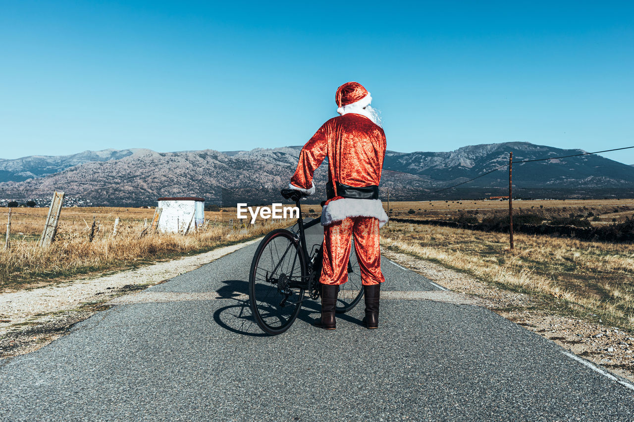 Back view of unrecognizable santa claus in red clothes standing with modern bicycle on empty road and looking away
