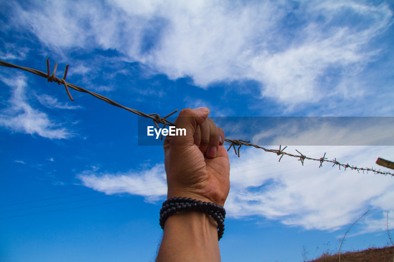 Cropped hand of man holding barbed wire against blue sky