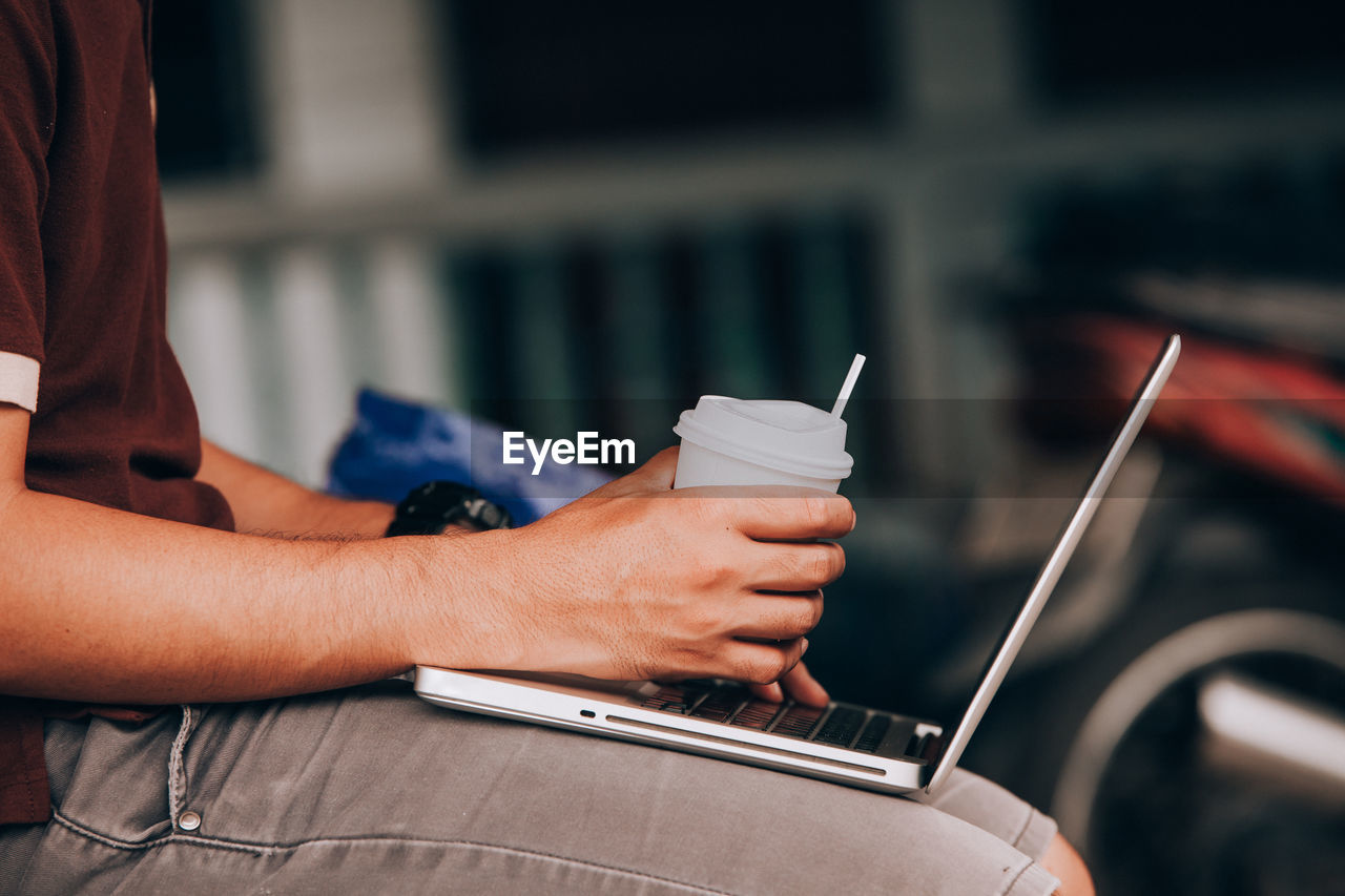 Cropped image of businessman holding disposable cup while using laptop