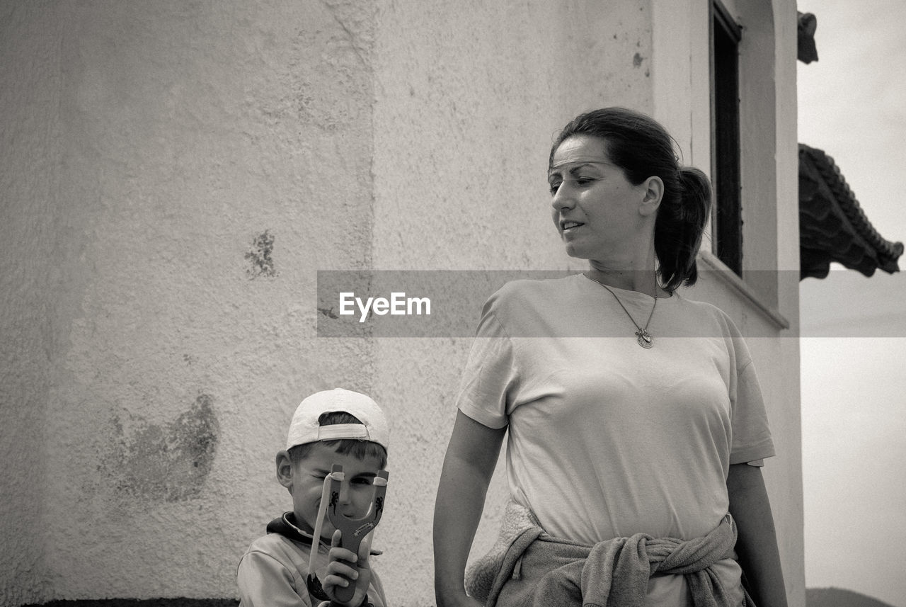 Portrait of boy looking through slingshot by mother against built structure