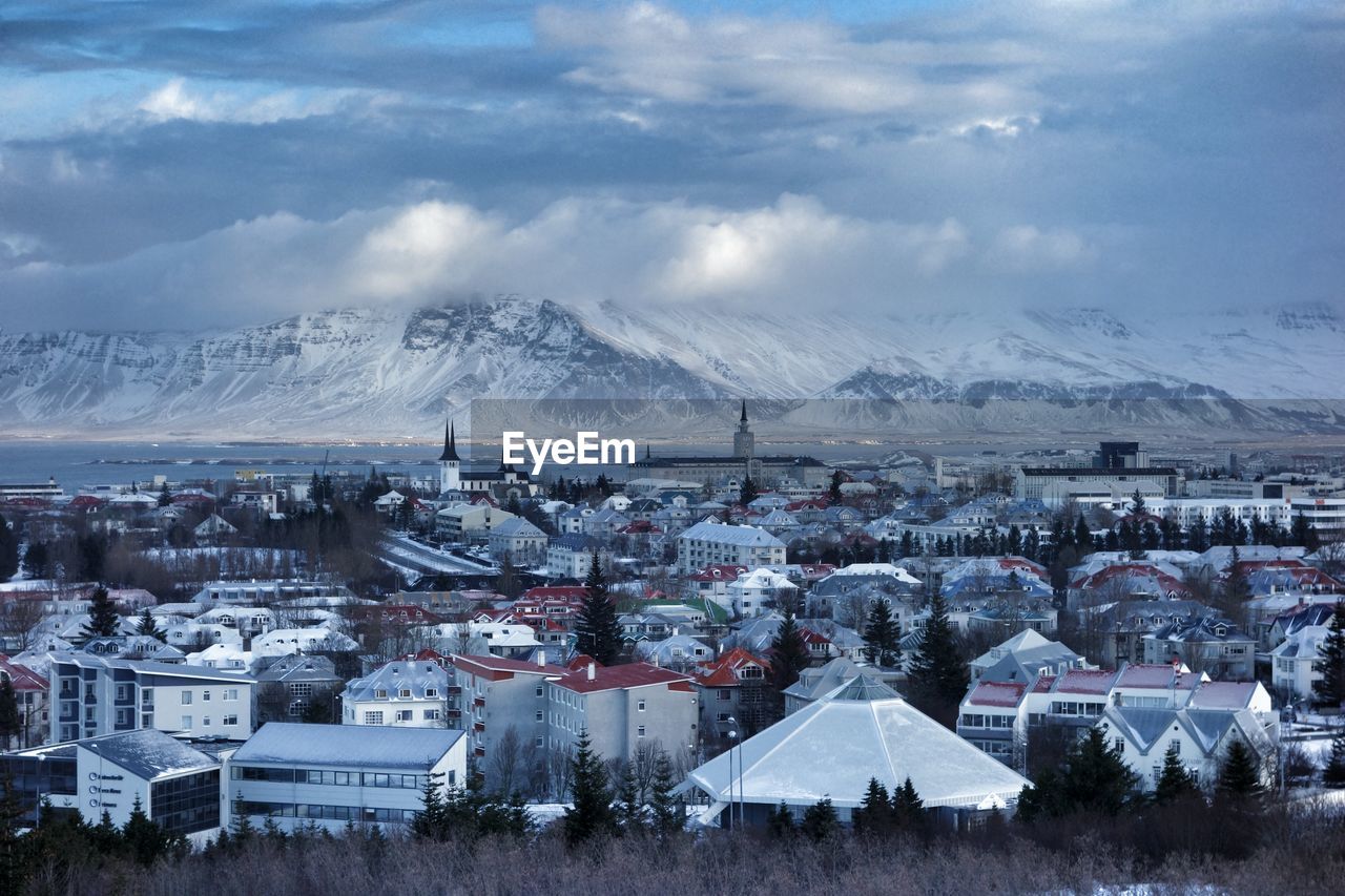 Buildings and snowcapped mountains against cloudy sky