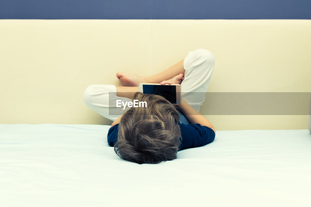 REAR VIEW OF BOY USING MOBILE PHONE WHILE ON BED