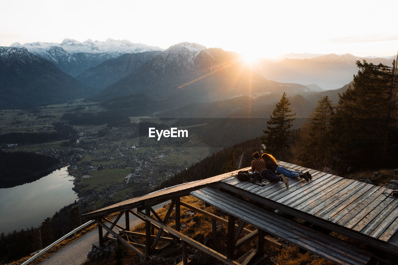 Back view of unrecognizable couple of travelers lying on wooden terrace and enjoying sunrise in highlands in foggy morning