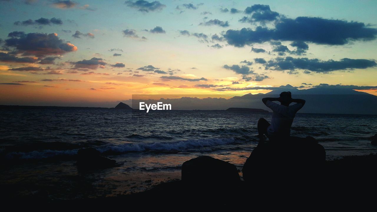 Silhouette man sitting on rock at beach against sky during sunset