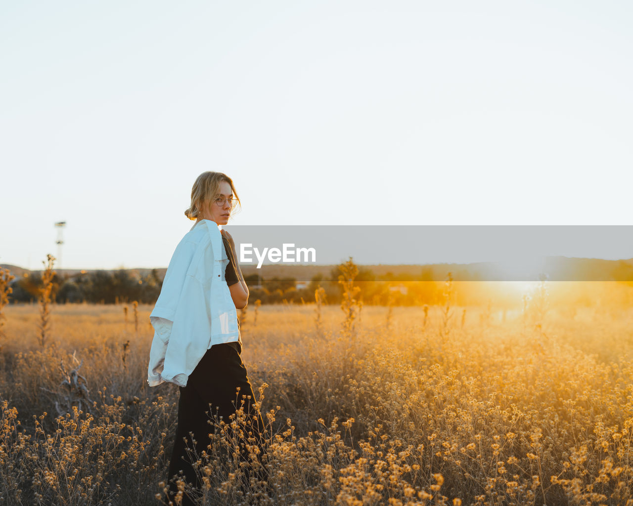 Side vie of trendy young female tourist with blond hair in casual clothes and eyeglasses walking on rural road amidst dry plants and looking over shoulder in countryside