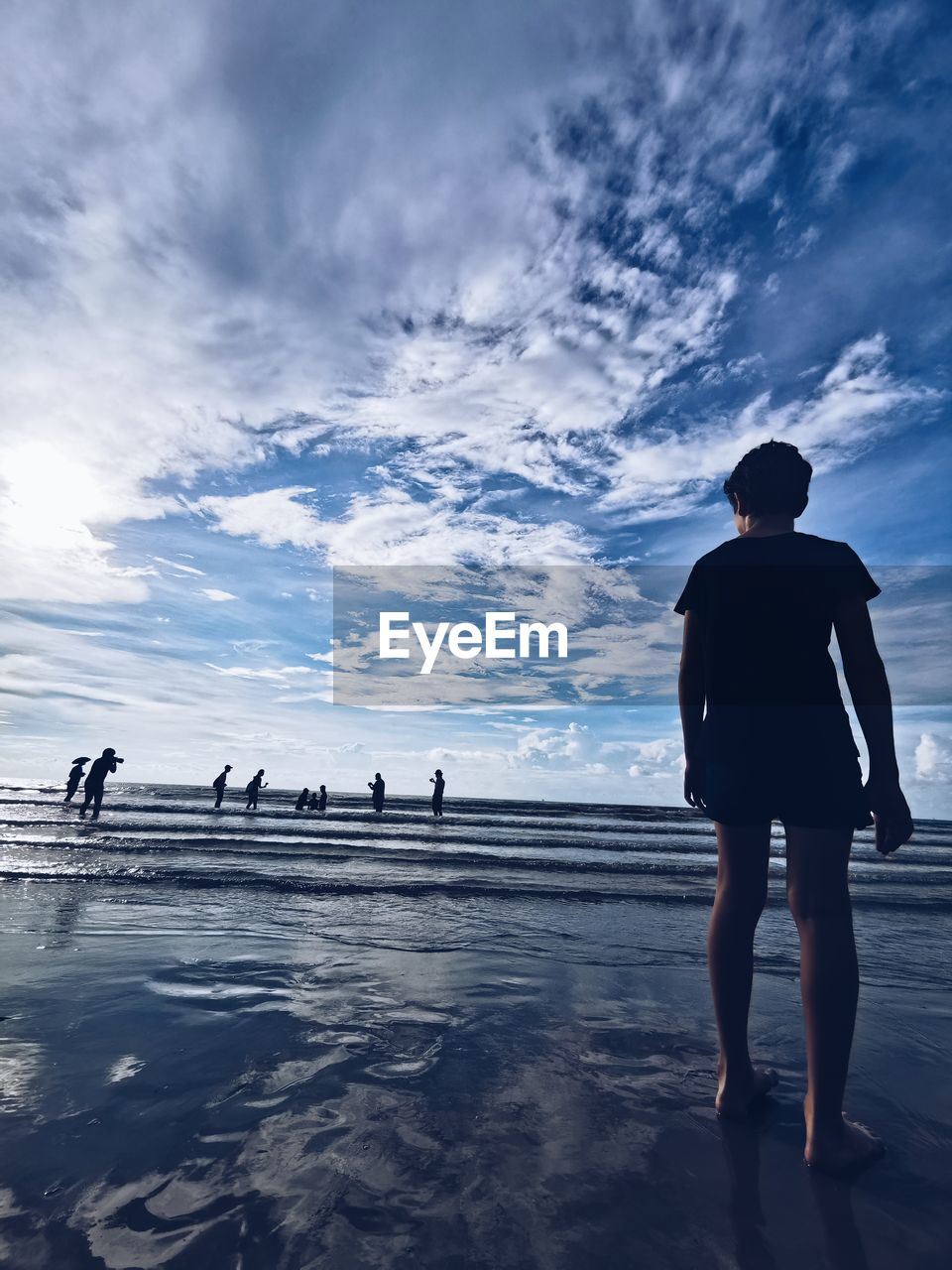 REAR VIEW OF MAN STANDING AT BEACH AGAINST SKY