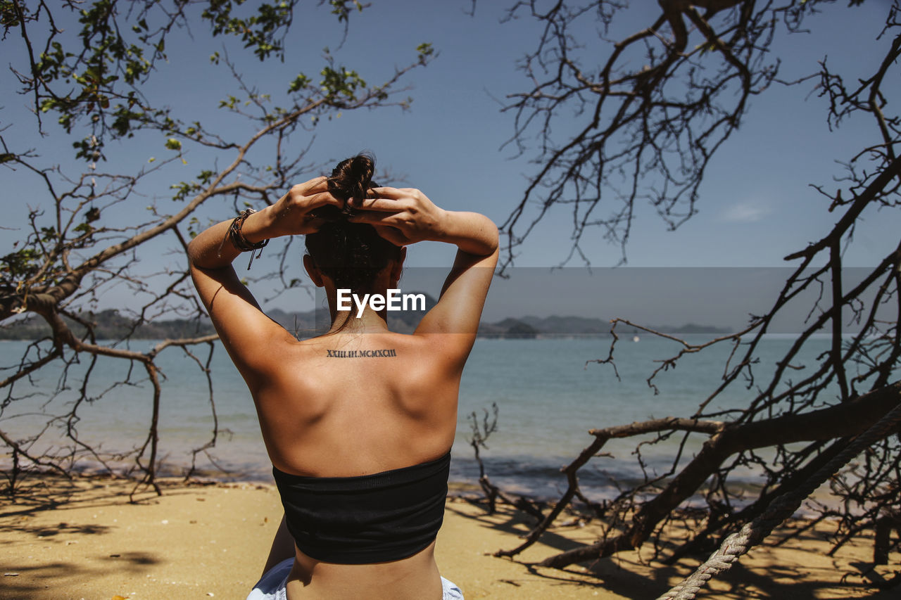 Rear view of woman tying her hair at beach against sky