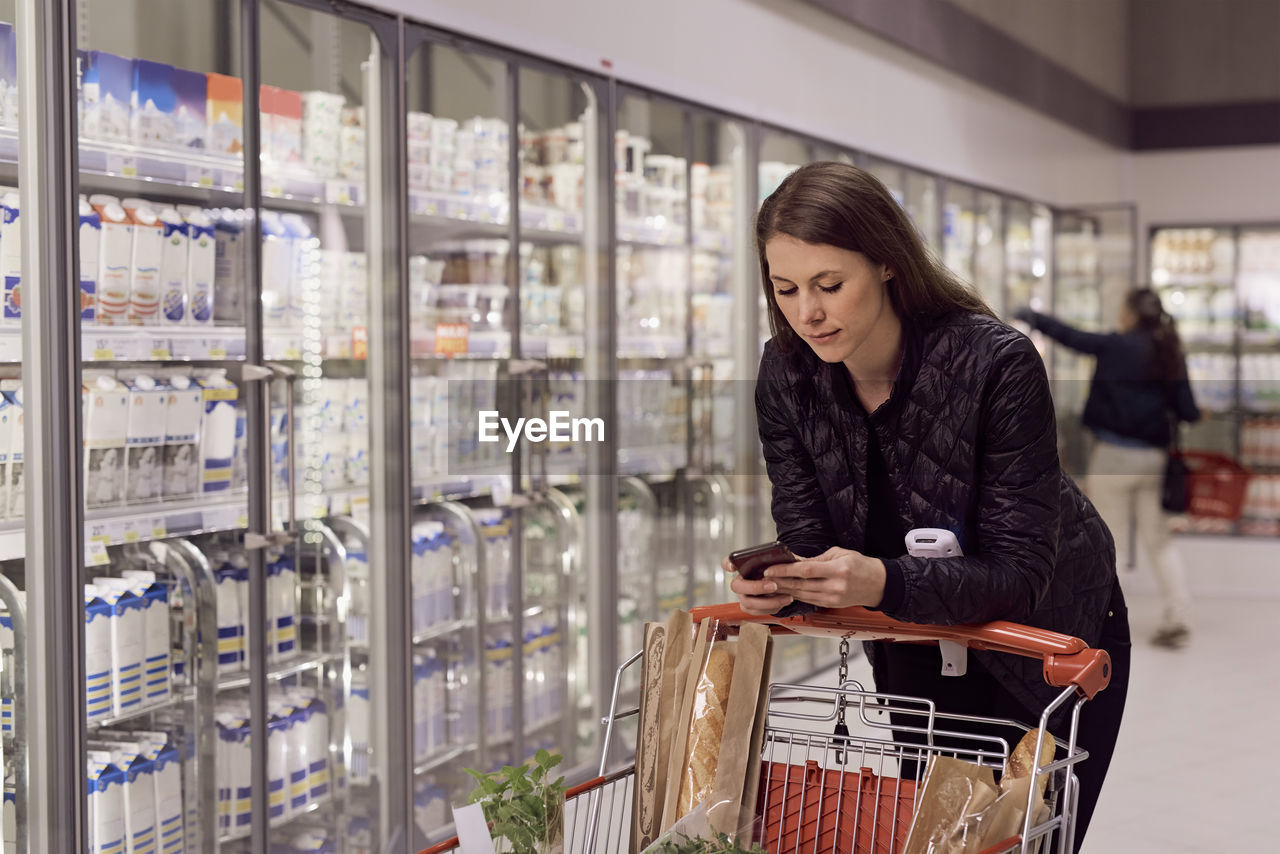 Woman text messaging while leaning on shopping cart at refrigerated section in supermarket